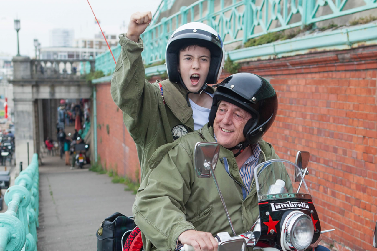 Two male characters are on a scooter, one of them has his hands up in the sky, cheering.